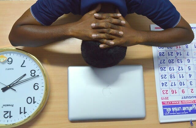  En övertrött man lägger huvudet på sitt skrivbord, bredvid sin laptop, kalender och klocka.